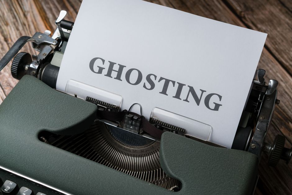 A vintage typewriter with a sheet of paper inserted, displaying the word 'GHOSTING' in bold, uppercase letters. The typewriter is placed on a wooden surface, showing its black and grey metal parts, including the roller, carriage, and keys. The background features additional blurred furniture and moving boxes, suggesting a home relocation or packing and moving process, which relates to house removals and transport services. The image emphasizes the concept of 'ghosting' in communication, situated within a setting of packing materials and moving equipment, indicating an ongoing or preparatory phase of a house removal carried out by Hounslow Removals.