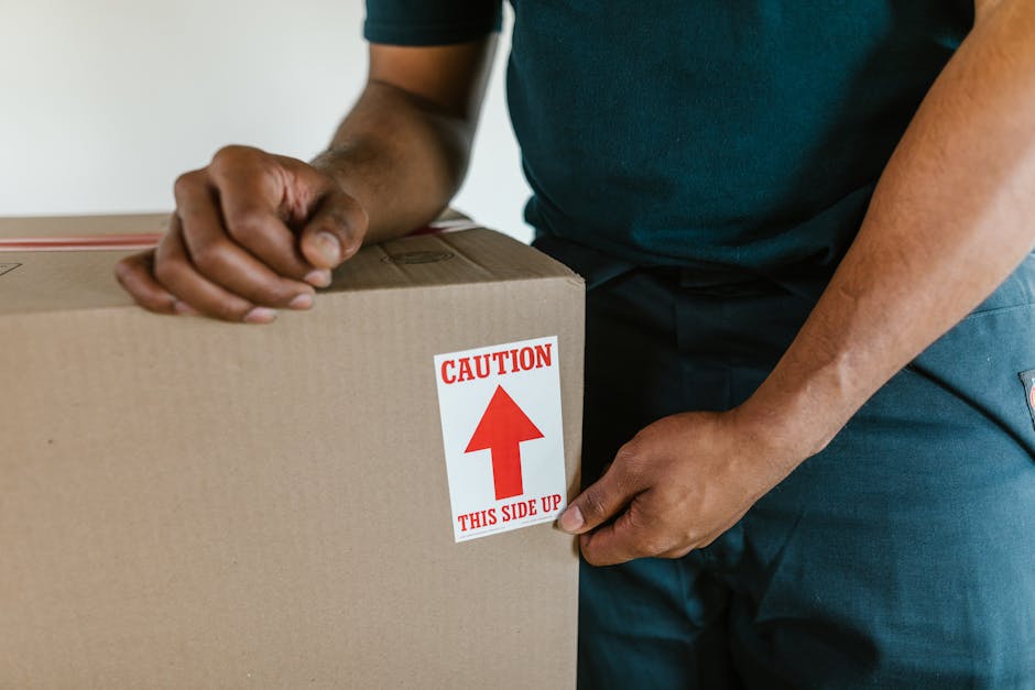 Close-up of a person in a dark shirt and blue trousers carefully handling a large cardboard moving box, which has a red and white caution sticker indicating 'Caution: This Side Up' with an upward pointing arrow, during a home relocation process. The person's hands are gripping the box firmly, with one hand on the top edge and the other supporting the side. The background shows an indoor setting with a neutral wall, and the scene captures the loading or packing phase of house removals, reflecting careful handling and proper packing materials used by [COMPANY_NAME] in furniture transport and packing and moving services. The box appears sturdy, made of brown kraft cardboard, suitable for protected transport of household items, and the overall environment suggests a professional move taking place inside a residential property.