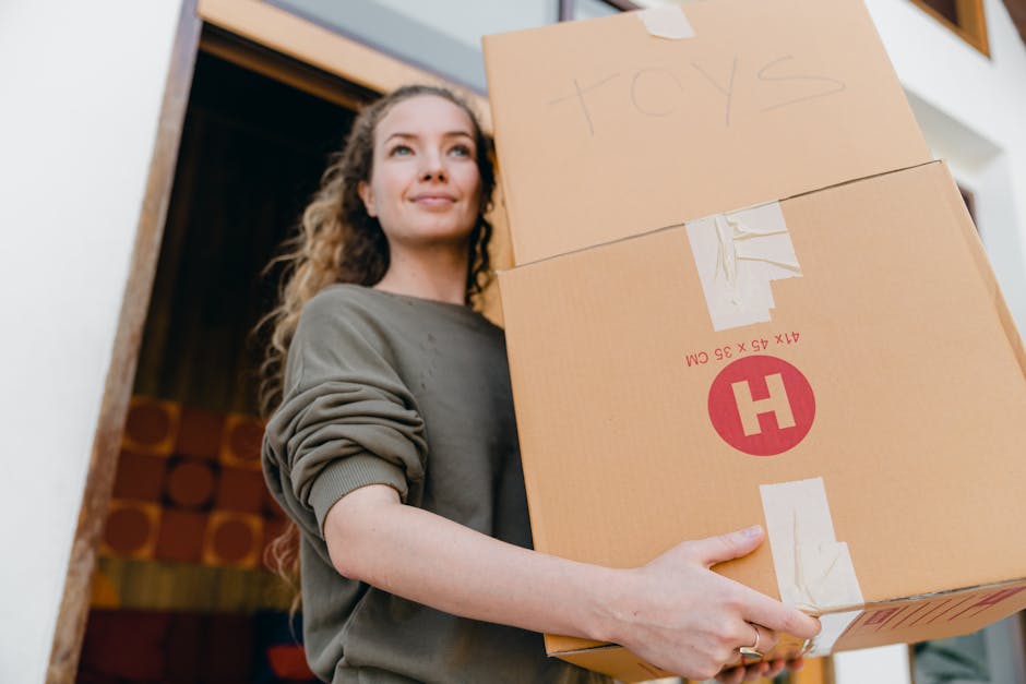 A young woman with long, curly hair and a neutral expression carrying two large cardboard boxes during a home relocation. One box is labeled 'toys' with handwritten text, while the other features printed red and white markings indicating the package weight and size. She is wearing a grey long-sleeve shirt with the sleeves rolled up and is positioned inside or near a doorway, with a staircase visible in the background. The boxes are wrapped with packing tape, and the woman is gripping them securely, demonstrating the process of packing and moving house. The environment suggests an indoor setting with natural lighting, and the image captures the loading process involved in professional removals services offered by Hounslow Removals, highlighting packing and furniture transport essentials.