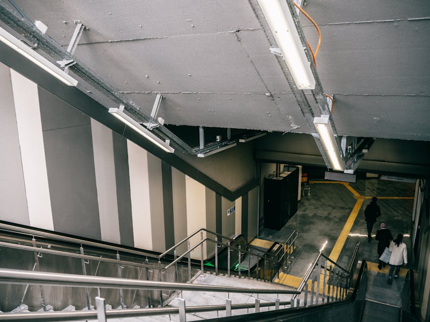 View from the top of a staircase inside a building leading down to Hounslow Central station, with three people walking along the platform. The staircase has metal handrails and concrete steps, while the ceiling above displays exposed electrical wiring and fluorescent lighting fixtures. The wall beside the stairs is painted with vertical grey and white stripes. The station's floor is made of grey tiles with yellow tactile paving strips for safety, guiding passengers toward station access. This scene is associated with moving and home relocation services, illustrating the logistics of stairs access during furniture transport, possibly as part of a home or office removal process involving Hounslow Removals.