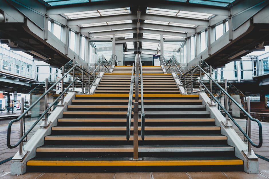 A wide, upward view of a modern, well-lit structure of a staircase leading to a train station platform, featuring black and yellow safety strips on each of the concrete steps with wooden treads. The staircase has metal handrails on both sides, and the structure is enclosed by glass panels and a metal framework, allowing natural light to illuminate the area. The surrounding environment includes part of the station's outer area visible through the glass, with some signage and urban elements in the background. This setting is suitable for illustrating the logistics involved in house removals and relocating items to the station, as managed by Hounslow Removals, with consideration of the staging and movement of furniture and packing materials during a home relocation process at Hounslow Central station.