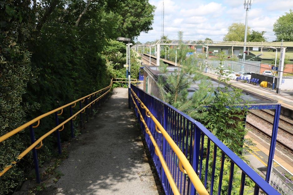 View from the top of a staircase inside a building leading down to Hounslow Central station, with three people walking along the platform. The staircase has metal handrails and concrete steps, while the ceiling above displays exposed electrical wiring and fluorescent lighting fixtures. The wall beside the stairs is painted with vertical grey and white stripes. The station's floor is made of grey tiles with yellow tactile paving strips for safety, guiding passengers toward station access. This scene is associated with moving and home relocation services, illustrating the logistics of stairs access during furniture transport, possibly as part of a home or office removal process involving Hounslow Removals.