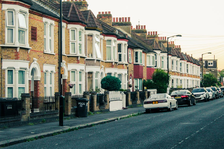 A clear street view on Lampton Road in Hounslow showing parked cars lining both sides of the road. On the left side, there are pedestrians walking on the sidewalk, with a woman carrying shopping bags and others near outdoor seating areas outside a pub or restaurant, which features black and white signage and large window displays. The sidewalk has large planters with colorful flowers, and a lamppost can be seen amongst the greenery. On the right side, the sidewalk is narrower with several vehicles parked close to the building facades, which include shops and a pharmacy with green signage. A large tree with lush green foliage extends over parts of the street, providing some shade. The road itself is empty of moving traffic, with a central dashed white line marking the lane, and the scene appears to be daytime with overcast lighting. This setting illustrates a typical urban environment suitable for house removals and furniture transport activities conducted by Hounslow Removals, emphasizing street access, parking considerations, and urban logistics involved in home relocation or packing and moving tasks.