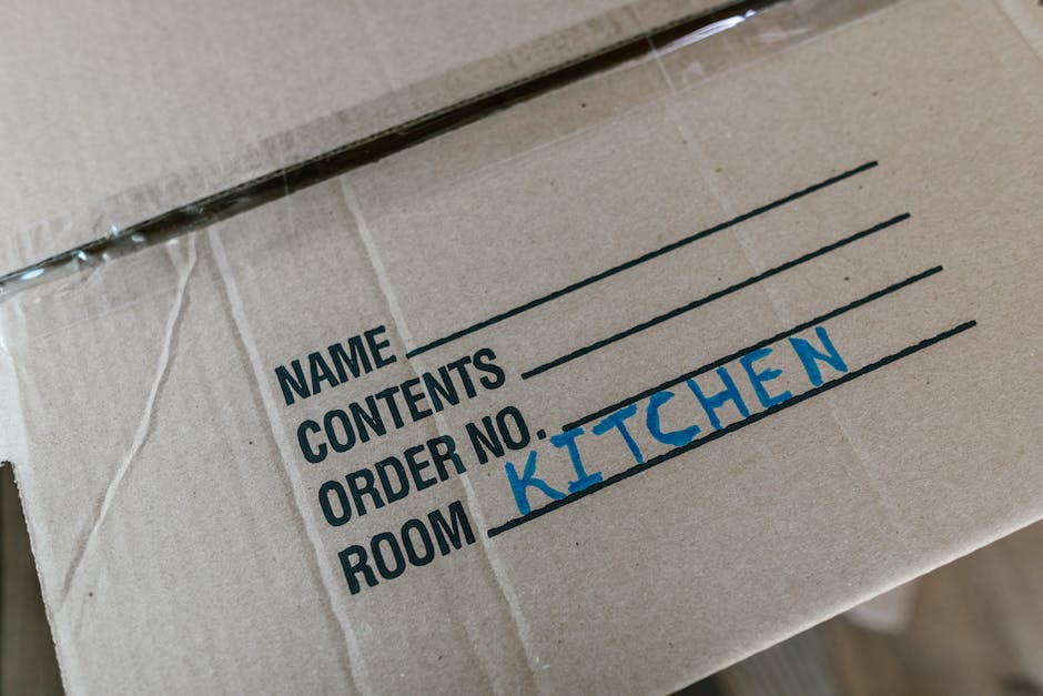 Close-up of a cardboard moving box with printed labels, showing handwritten text indicating it belongs to the kitchen. The box is secured with clear packing tape and is situated inside a home, likely in the entrance or hallway, ready for loading during a house removal. Other boxes and packing materials, such as bubble wrap or tissue paper, may be visible nearby, along with furniture or appliances in the background. The environment appears well-lit, possibly from natural light through windows. This image reflects the packing and logistics involved in a home relocation, as handled by professional removals services like Hounslow Removals, which support efficient furniture transport and moving processes.