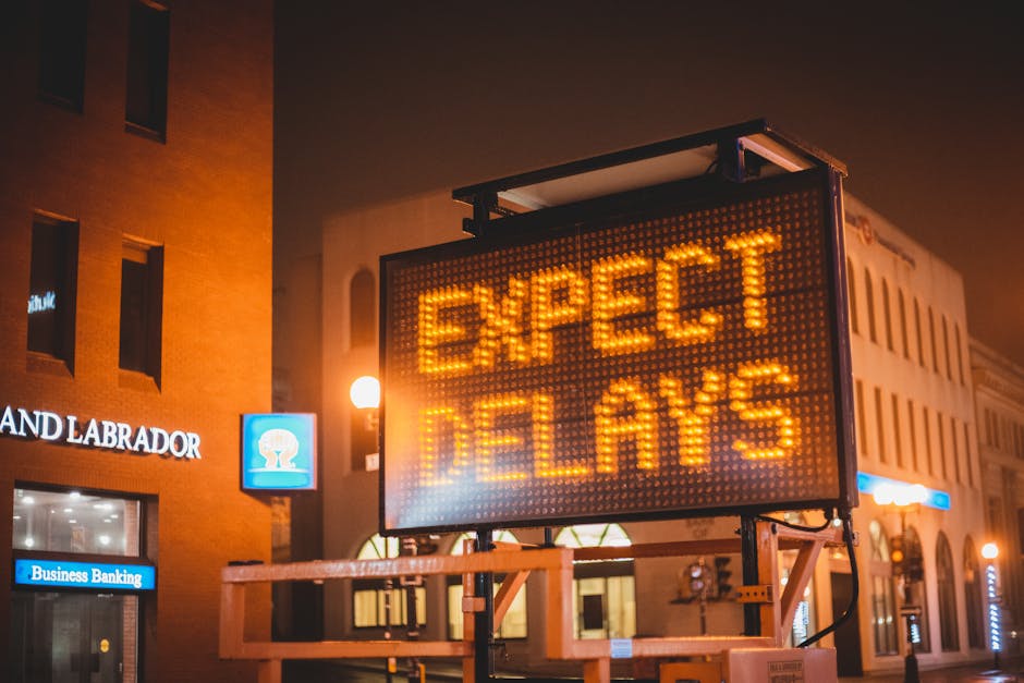 A large electronic road sign mounted on a stand displayed at night, with illuminated orange-yellow text reading 'EXPECT DELAYS' in a busy urban area. The sign is positioned on the pavement in front of a multi-storey building with lit windows and signage, including a blue Business Banking sign and a white sign reading 'LAND LABRADOR.' The surrounding buildings are constructed from brick and have architectural details typical of a city centre. Street lamps cast a warm glow over the scene, and the sign appears to be part of a traffic management or public information system related to road congestion or ongoing work, relevant to home relocation or moving logistics as discussed on the webpage about same-day removals delays.