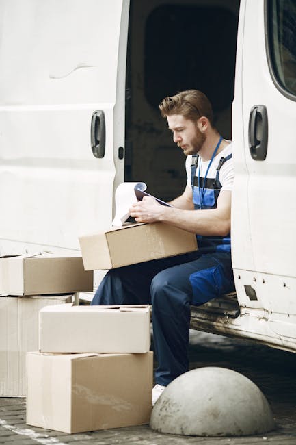 A male mover with light skin, short brown hair, and a beard, is seated on the edge of a white delivery van with its sliding door open, during a home relocation process. He is wearing a striped t-shirt and blue work overalls, holding a clipboard and pen, and appears focused on taking notes. Surrounding him are several cardboard boxes of various sizes, some stacked on the ground and one resting on his lap, which are typical packing materials used in removals and furniture transport. The van is parked on a paved surface, with a concrete bollard nearby, indicating an outdoor setting close to a property. Inside the van, additional packing and loading materials are visible, supporting the logistics involved in packing and moving tasks. The environment suggests early stage of a house removal or similar service, with attention to organized packing and strategic loading, associated with professional removals services offered by Hounslow Removals.