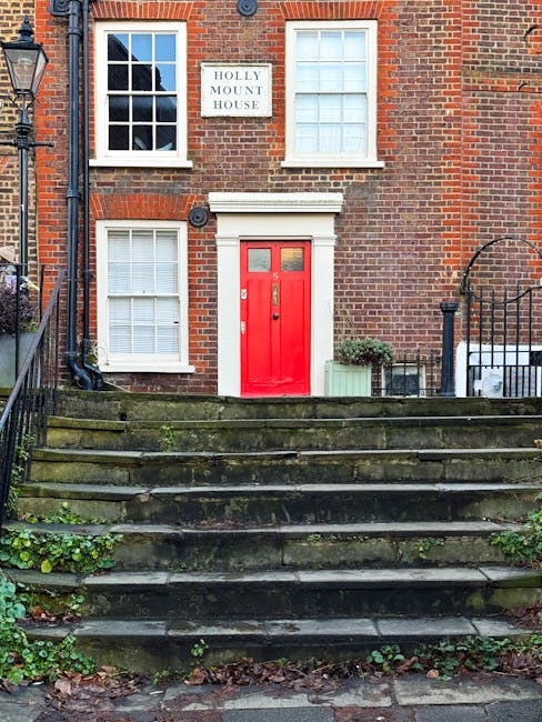 The image shows the front of a brick residential building with a red door and white-framed windows on the first and second floors. A small sign above the door reads 'Holly Mount House.' The building is accessed via a set of stone steps with some moss and greenery growing between them, leading up to the entrance. To the right of the door, there is a green plant in a rectangular planter box, and a black iron fence with a gate partially visible, indicating an enclosed front area. The overall scene captures the exterior of a typical UK housing property, relevant to house removals and moving services, with natural daylight illuminating the scene.