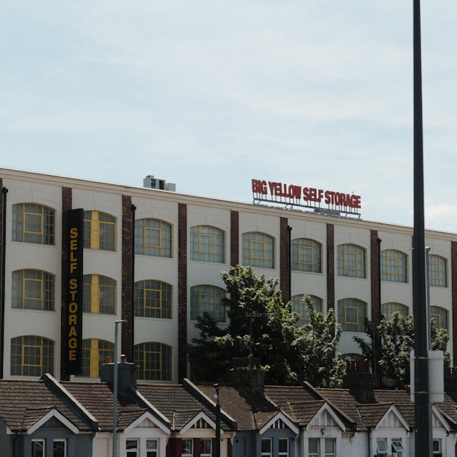 A multi-storey commercial building with a white facade and large, arched windows featuring yellow frames, situated behind a row of small residential houses with pitched roofs and chimneys. The building displays a vertical sign indicating 'Self Storage' in black letters on a yellow background, and a red sign on the roof reading 'Big Yellow Self Storage'. The scene is set during daytime with a partly cloudy sky and sunlight illuminating the structures. In the foreground, a streetlamp and a utility pole are visible, with some trees partially obscuring the lower part of the commercial building. The image captures the typical environment surrounding a self-storage facility, highlighting the urban setting where home relocation and furniture transport services by Hounslow Removals might operate, especially in properties with limited access requiring specialist loading solutions.