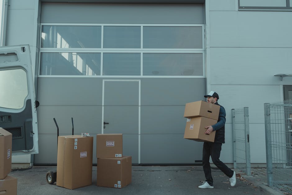 A man wearing a black baseball cap, black jacket, and dark trousers is carrying two cardboard boxes, one on top of the other, during a home relocation process outside a modern industrial building with grey metal paneling and large glass windows. Several packing boxes of various sizes, some with labels and symbols, are arranged on the pavement nearby. Two hand trucks with upright handles are positioned on the ground, suggesting they are being used to facilitate the loading and unloading of items. The van's rear door is open, indicating it is being loaded with moving boxes and furniture as part of a professional removals service, likely connected to house moving or furniture transport. The surroundings include part of a metal fence, and the scene is lit by natural daylight with a clear focus on the moving activity, illustrating a typical packing and loading process for an upcoming house relocation, as outlined in Hounslow Removals' TW3 to TW5 moving tips.