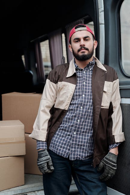 A young man with a beard and wearing a red cap backward, a checked shirt, and a beige and brown jacket stands inside a van during a house move, with cardboard boxes stacked behind him on the vehicle's cargo area floor. The van's interior shows a dark ceiling and side panel, and the light coming in highlights the packing materials and the man's neutral expression. This scene illustrates packing and loading processes typical of home relocation or furniture transport services, as part of a professional removals operation by Hounslow Removals in a residential setting.