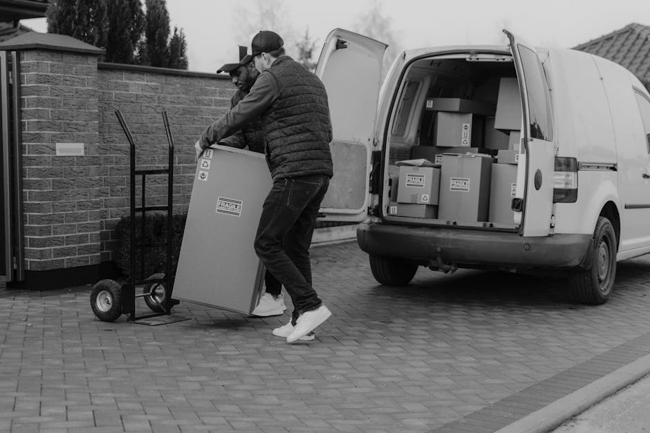A man wearing a black baseball cap, black jacket, and dark trousers is carrying two cardboard boxes, one on top of the other, during a home relocation process outside a modern industrial building with grey metal paneling and large glass windows. Several packing boxes of various sizes, some with labels and symbols, are arranged on the pavement nearby. Two hand trucks with upright handles are positioned on the ground, suggesting they are being used to facilitate the loading and unloading of items. The van's rear door is open, indicating it is being loaded with moving boxes and furniture as part of a professional removals service, likely connected to house moving or furniture transport. The surroundings include part of a metal fence, and the scene is lit by natural daylight with a clear focus on the moving activity, illustrating a typical packing and loading process for an upcoming house relocation, as outlined in Hounslow Removals' TW3 to TW5 moving tips.